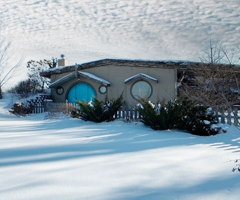 Winter scape of the "Hobbit" style Cove cabin with a round blue door, evergreen shrubs, and long shadows in the snow.