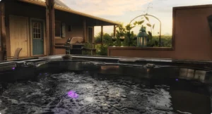 An idyllic vacation rental cabin features an outdoor hot tub on a wooden deck, overlooking a charming house. Enjoy the grill, a table set with two wine glasses, and a lantern—all framed by a scenic sunset backdrop for perfect relaxation.