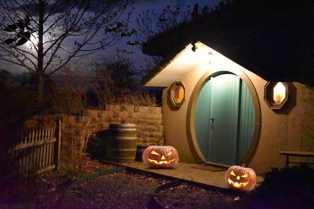 Hobbit style cabin with round blue door, two jack-o-lanterns, bats and witches in an autumn sky.