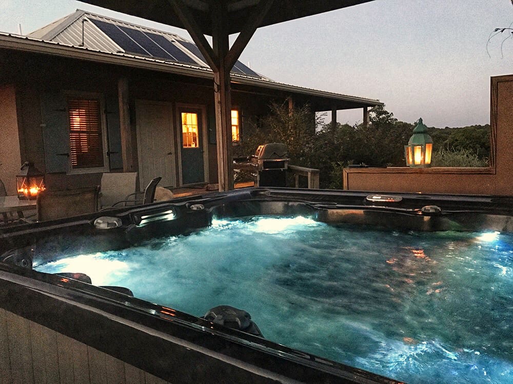 Large hot tub in the evening with blue water and the glow of cabin lights from inside.